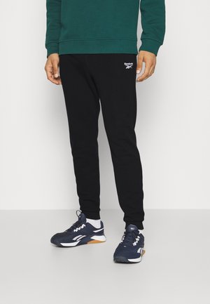 Man die zwarte Reebok joggingbroeken, donkerblauwe en witte Reebok sneakers en een groen sweatshirt draagt, staand op een lichtgrijze vloer.