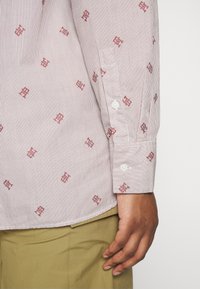 Camicia a maniche lunghe a righe bianca e rossa con piccoli loghi ricamati. Presenta polsini con bottoni e orlo piegato con una texture liscia.
