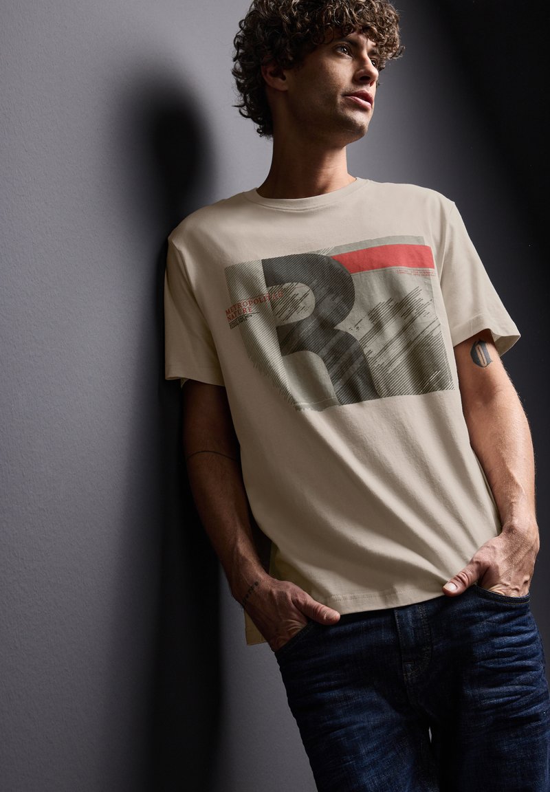 Ung man med lockigt hår som lutar sig mot en vägg, klädd i en beige t-shirt med tryck och mörkblå jeans, med händerna i fickorna och en sidoblick.