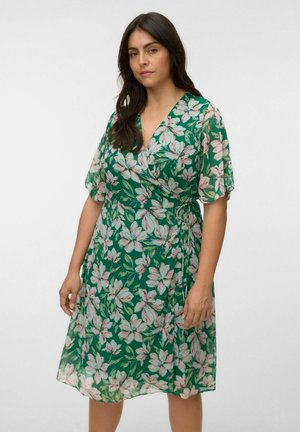 Kvinde med langt mørkt hår iført en knælængde grøn blomsterprint omslagskjole med korte ærmer, stående mod en ensfarvet baggrund.