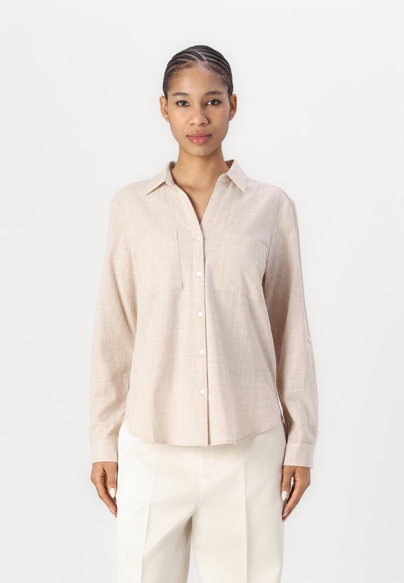 Beige button-up shirt with a textured pattern, long sleeves, and two chest pockets, paired with light-colored wide-leg trousers.