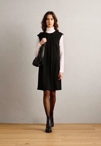 Vestido negro sin mangas hecho de una tela suave, con un corte holgado. Llevado sobre un suéter de cuello alto blanco, complementado con botas negras hasta el tobillo y un bolso a rayas.