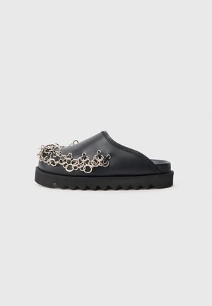 Chaussure noire à enfiler avec une semelle épaisse, présentant un design de chaîne en argent décoratif sur le dessus, texture lisse et semelle en zigzag.