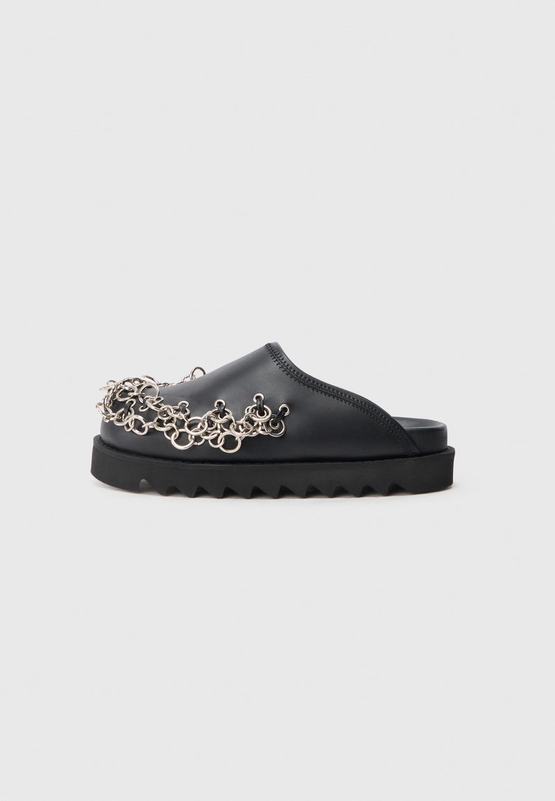Černá slip-on obuv s masivní podrážkou, která má dekorativní stříbrný řetěz na svršku, hladkou texturu a vlnitou podrážku.