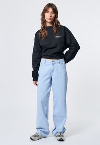 Schwarzer, kurzer Sweatshirt mit weißem Text und hellblauen weiten Jeans. Modell trägt graue und schwarze Sneakers und steht vor einem neutralen Hintergrund.