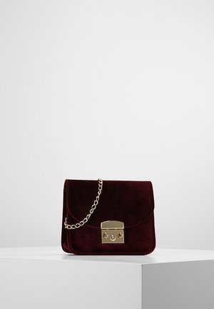 Sac à main en velours bordeaux foncé avec fermoir en métal doré et bandoulière chaîne dorée, présenté sur un piédestal blanc devant un fond gris.