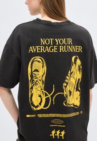 Μαύρο μπλουζάκι με κίτρινο σχέδιο από δένοντας κορδόνια αθλητικών παπουτσιών και το κείμενο «ΔΕΝ ΕΙΣΑΙ Ο ΜΕΣΟΣ ΔΡΟΜΕΑΣ» στην πίσω πλευρά, φορεμένο από ένα πρόσωπο με μακριά μαλλιά.