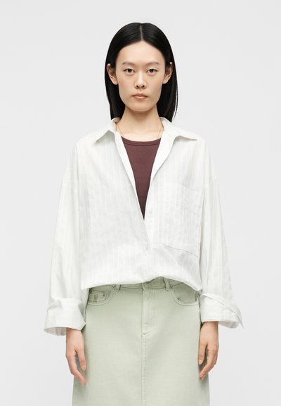 Femme aux cheveux noirs et raides porte une chemise blanche ample à boutons par-dessus un haut marron et une jupe vert clair, debout devant un fond uni.