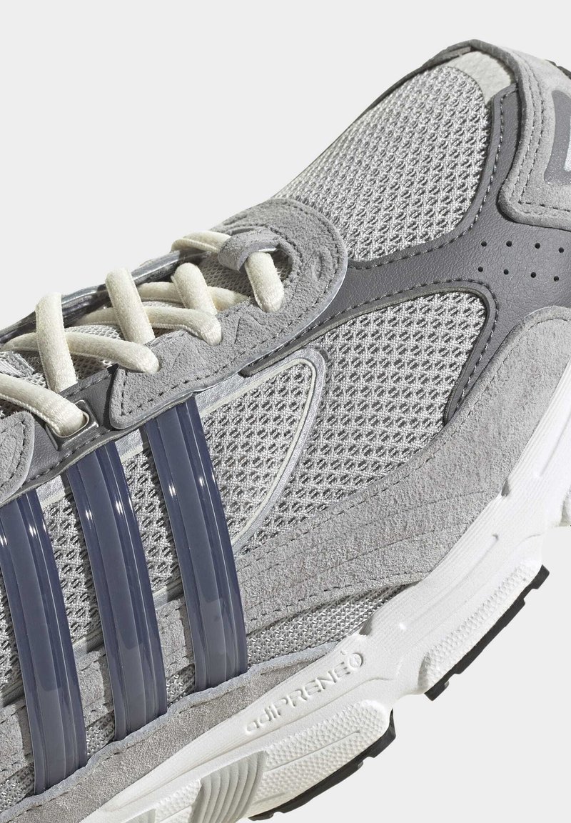 Zapatilla de running de tejido gris con parte superior de malla, diseño de plástico gris oscuro en forma de tres barras, cordones blancos y suela de goma texturizada.