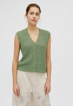 Joven mujer con un chaleco sin mangas verde de punto con botones y pantalones sueltos blancos de encaje con flores, de pie frente a un fondo liso.