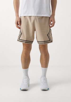 Hombre con pantalones cortos deportivos beige con rayas angulares negras, calcetines deportivos blancos y zapatillas blancas, de pie frente a un fondo blanco liso.