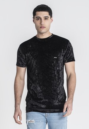 Camiseta de terciopelo negro con cuello redondo y mangas cortas; cuenta con una pequeña etiqueta texturizada en el pecho. Combinada con jeans de denim claro desgastados.