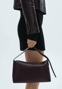 Sac à main en cuir bordeaux avec une texture lisse, de forme rectangulaire et une bandoulière amovible. Fermeture zippée sur le dessus.