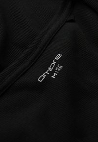 Zwarte stof met ribbelstructuur, voorzien van een witte gedrukt label met de tekst "Ombre M 148." De nadruk ligt op het materiaal en het ontwerp van het label.