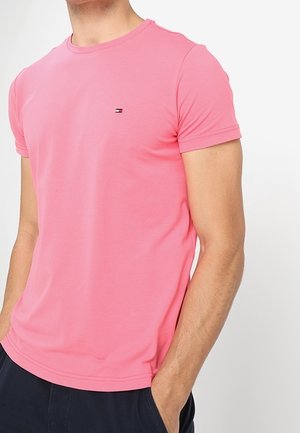 Hombre que lleva una camiseta rosa lisa de manga corta con un pequeño logo en el pecho y pantalones oscuros con una mano en el bolsillo.