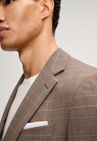 Brązowa marynarka w kratę o zorganizowanym kroju, z pomarańczowymi akcentami, klapami oraz kieszenią na piersi, w której znajduje się biały chusteczka.