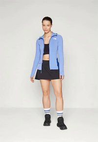 Γυναίκα στέκεται φορώντας ένα ανοιχτό μπλε φερμουάρ πανωφόρι, μαύρο κροπ τοπ, μαύρη φούστα, ριγέ κάλτσες και μαύρες ορειβατικές μπότες σε απλό φόντο.