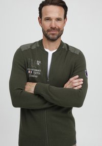 Zelený pletený kardigán so zipsom, kontrastnými ramennými nášivkami, výšivkou loga a ribbovanými manžetami a lemom.