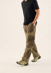 Olive green cargo pants, z wieloma kieszeniami i różnorodnymi fakturami. W połączeniu z czarną koszulką i lekkimi, wzorzystymi butami.