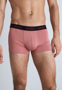 Hugo vīriešu bokseršortus gaiši rozā audumā ar melnu elastīgu jostasvietu, uz kuras redzams zīmola nosaukums sarkanā krāsā. Slim fit un gluda tekstūra.