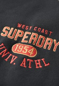 Sort stof med rødt og guld broderet tekst: "WEST COAST SUPERDAY 1954 UNIV. ATHL." Glat tekstur med kontrasterende farver.