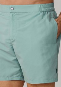 Bañadores de hombre de color verde claro con cintura elástica, cierre de botón y bolsillos laterales, usados por una persona con la mano en el bolsillo.