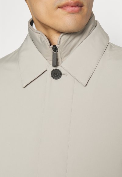 Chaqueta ligera beige con cuello alto, cierre de botones negros y detalles de cremallera, hecha de un tejido suave con costuras sutiles.