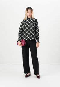 Donna in piedi che indossa una blusa a fantasia bianco e nero, pantaloni neri, scarpe bordeaux e tiene una pochette bordeaux coordinata in un contesto bianco.