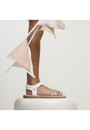 Sandal de cuero blanco con correa de hebilla, que presenta un acento decorativo dorado en la punta, posicionado sobre un pedestal de color claro.