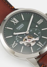 Rund Fossil automatisk armbåndsur med grøn urskive, sølv romertal, eksponerede mekaniske tandhjul og brun læderrem. Klokken viser 12:09.