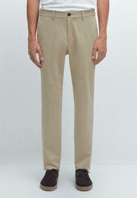 Pantalon beige en tissu lisse, avec une coupe droite, une poche avant unique et une fermeture à bouton. Porté avec des chaussures foncées.