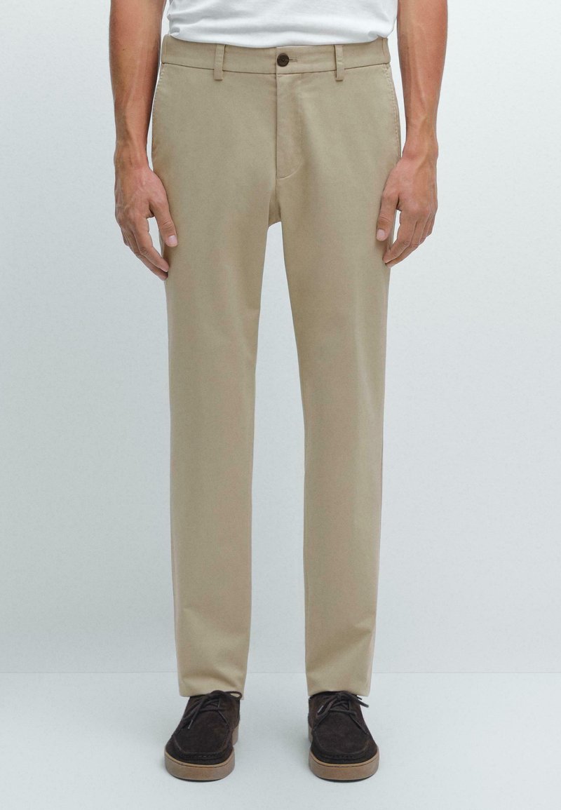 Pantalon beige en tissu lisse, avec une coupe droite, une poche avant unique et une fermeture à bouton. Porté avec des chaussures foncées.