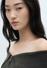 Model met donker, steil haar die een donkere off-the-shoulder top met een ritssluiting draagt. Gladde huid met neutrale tinten, minimale make-up.