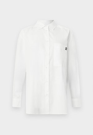 Cameră albă cu mânecă lungă, confecționată dintr-un amestec de bumbac, având un guler cu nasturi, un buzunar în față și un mic logo negru pe piept.