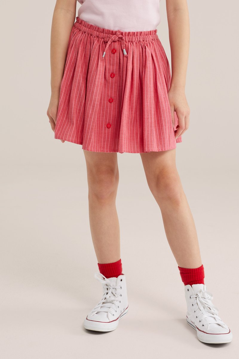 Kind trägt einen rosa Rock mit vertikalen Streifen und roten Knöpfen, rote Socken und weiße High-Top-Sneaker, steht vor einem beigen Hintergrund.