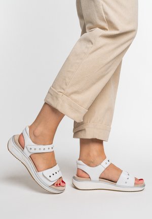 Feet wearing white sandals with silver studs and beige rolled-up corduroy pants against a plain white background.