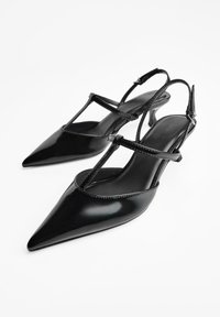 Čierne patentované kožené slingback opätky so špicatými prstami, s jemnými popruhmi a nízkym, zužujúcim sa opätkom. Hladká textúra s lesklým povrchom.