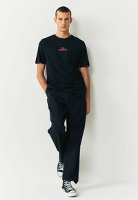 Melnas kokvilnas T-krekls ar uzrakstu "THE EXORCIST" sarkanā krāsā, apvienots ar brīviem melniem biksēm un melniem augstiem kedām.