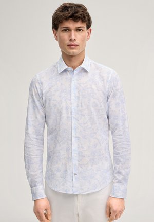 Junger Mann mit lockigem Haar, trägt ein hellblaues, langärmliges Hemd mit Blumenmuster, steht vor einem einfarbigen Hintergrund.