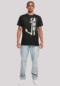 Black cotton t-shirt features bold white text and an anchor graphic, paired with light blue jeans and white sneakers.