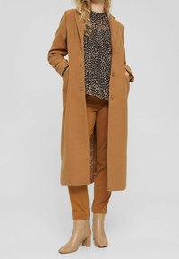 Cappotto lungo di cammello con due bottoni, blouse nera a fantasia e pantaloni marroni attillati. Stivali beige alla caviglia completano l'outfit.