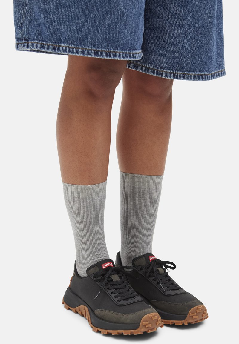 Gambe che indossano calze grigie fino al polpaccio, scarpe sportive nere e marroni con lacci e pantaloni corti in denim blu fino al ginocchio su sfondo bianco.