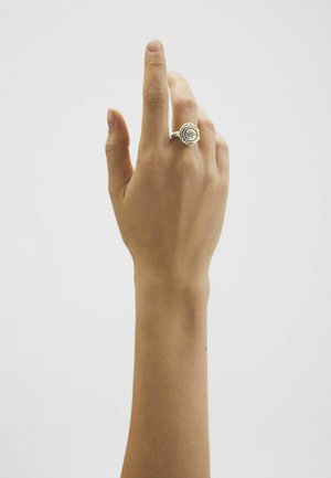 Labā roka ar paceltu vidējo pirkstu, uz vidējā pirksta valkājot sudraba spirāles dizaina gredzenu, uz vienkārša balta fona.