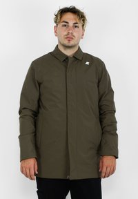 Cappotto impermeabile verde oliva con colletto alto, bottoni frontali e tasche laterali. Presenta un piccolo emblema sul lato sinistro del petto.