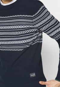 Marinblå stickad tröja med vita geometriska mönster, rundad halsringning, strukturerad tygkvalitet och en liten märkesetikett längst ner till vänster.