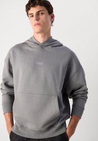 Grå hettegenser laget av myk stoff, med en frontlomme, ribbestrikkede mansjetter og en minimalistisk logo på brystet. Justerbar snorhette.