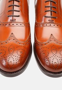 Scarpe da uomo in pelle marrone con design wingtip, caratterizzate da dettagli perforati, texture liscia e suola scura a contrasto.