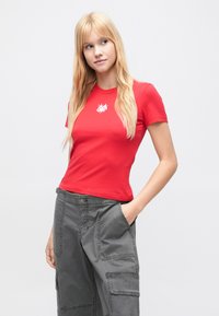 Νεαρή γυναίκα με μακριά ξανθά μαλλιά, φοράει κόκκινο t-shirt με μικρό λευκό σχέδιο και γκρι παντελόνι cargo, τα χέρια στις τσέπες, κοιτάζει πλάγια.