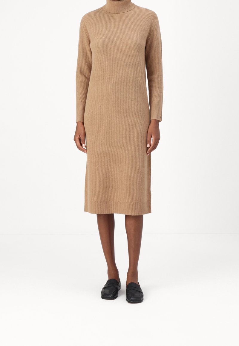 Robe en maille de couleur camel avec un col montant et des manches longues. Le design présente une coupe droite et une texture côtelée. Associée à des chaussures noires.