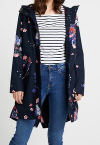 Marineblå blomstret regnfrakke med hætte, prydet med pink og hvide blomster, en knaplukning foran og lommer, båret over en stribet top og jeans.